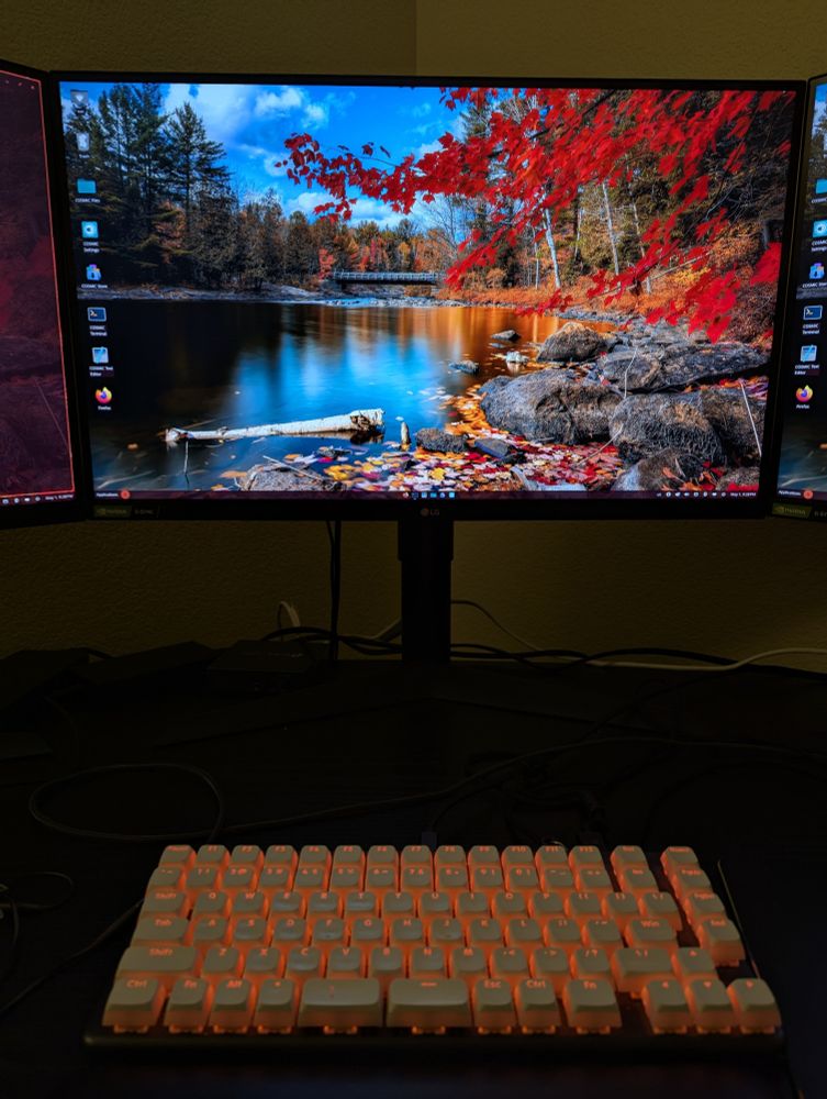 A picture of my desktop with my monitor at the top and my keyboard at the bottom. The monitor is showing the COSMIC desktop environment with a colorful background. The keyboard has LEDs that are matched to an accent color extracted from that background, in this case, orange.