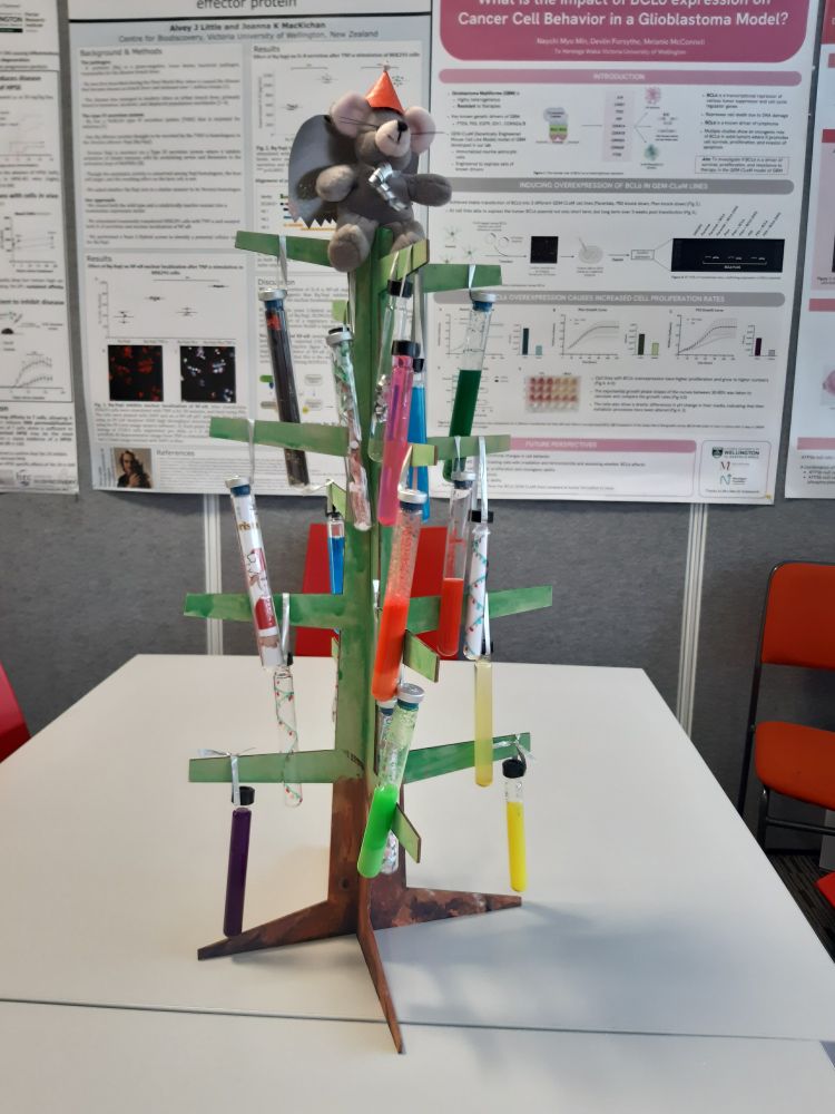 A small bare-bones wooden Christmas tree with a Christmas lab mouse/ angel on the top. It is decorated with hanging test tubes filled with a variety of contents!