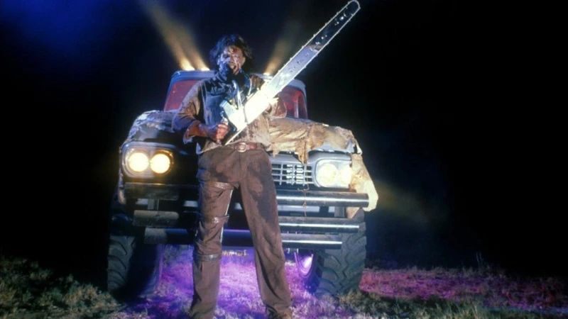 A film still from LEATHERFACE: TEXAS CHAINSAW MASSACRE III, depicting Leatherface standing in front of a pick up truck and holding a comically large chainsaw. 