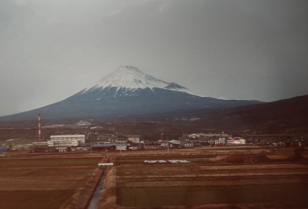 Mt Fuji in silhouette, capped with snow. The fields in front of the mountain are brown and a river flows out towards the viewpoint 
