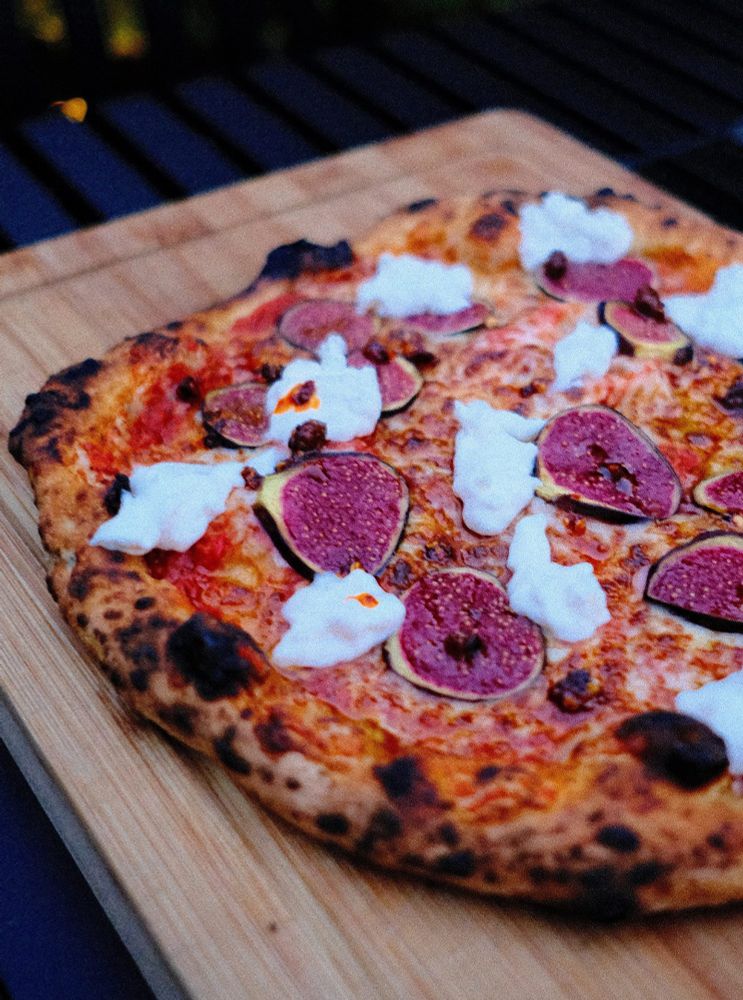A pizza sits on a wooden board. The pizza is topped with sliced figs and dollops of creamy white stracciatella cheese. The crust is nicely spotted 