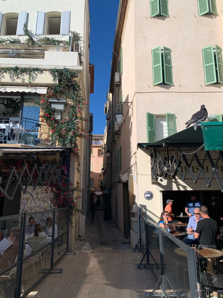 L’entrée d’une ruelle très étroite. La façade de droite, une terrasse de café, est au soleil, le reste est dans l’ombre.
