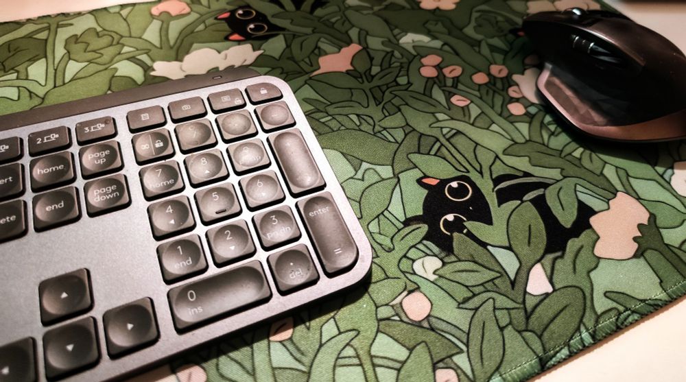 A desk mat with black cats peeking out from behind thick, leafy green foliage and flowers 