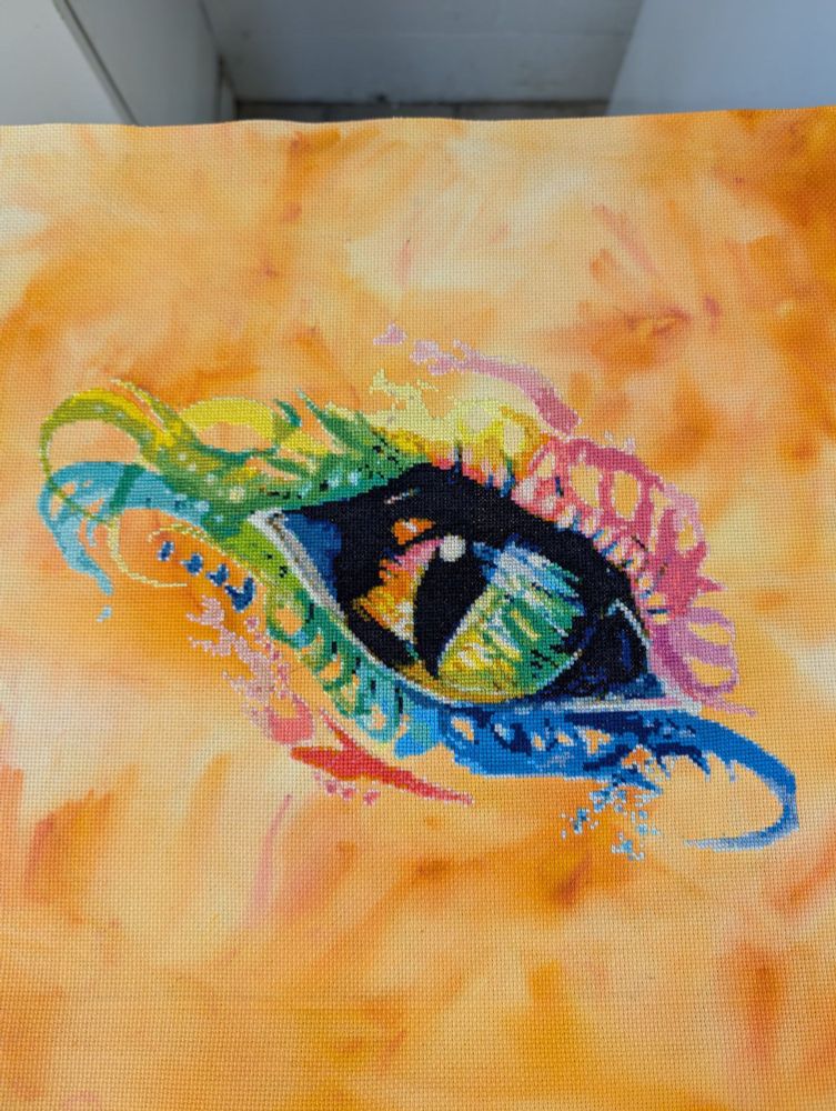 Cross stitch piece of a rainbow dragon eye on orange dyed cloth. Katy Lipscomb art that was made into a cross stitch pattern.