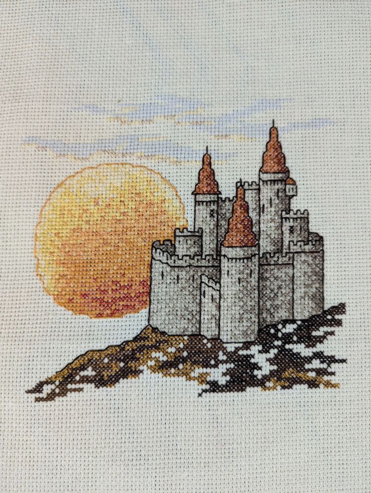 Cross stitch of a castle with the sun and some clouds on a beige Aida cloth. Back stitch details for castle and around the sun.