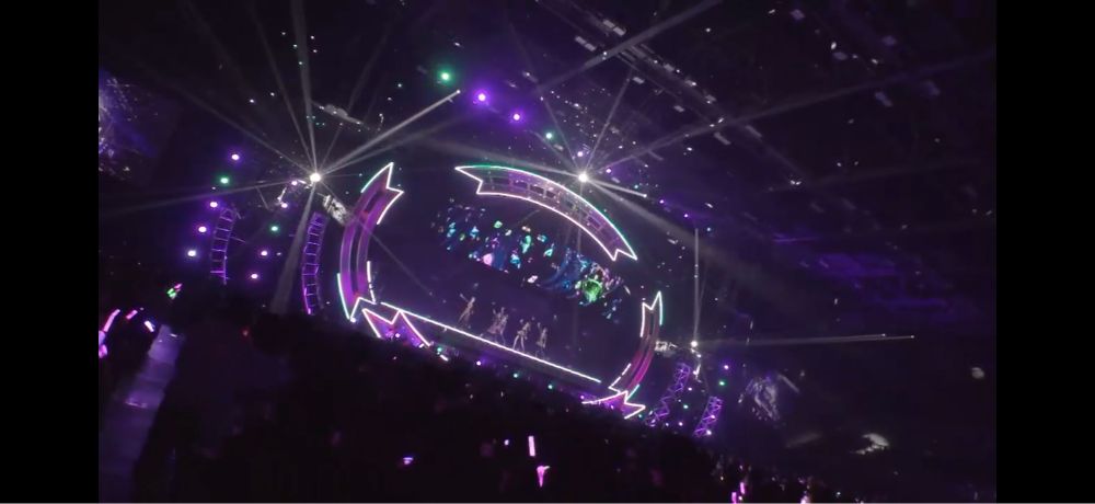 舞台照明の白、濃いピンク、緑といった色のライトやミラーボールによって、煌びやかに輝いているステージと、ステージを見つめる観客たちの様子。