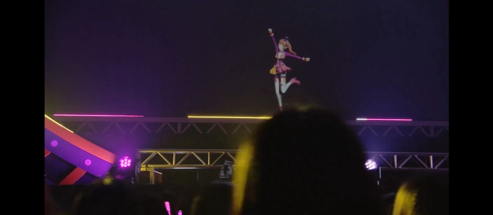 ステージ上で元気よく踊る水嶋咲。右手をあげ、左膝を後ろに曲げているような状態。
また、ステージを見る観客たちの後頭部も画像下部に見えている。