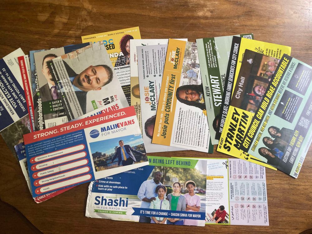 A pile of campaign flier mailers for Rochester mayor and city council spread out across a table.