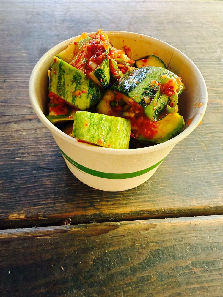 A paper cup of zucchini kimchee on a wooden table.