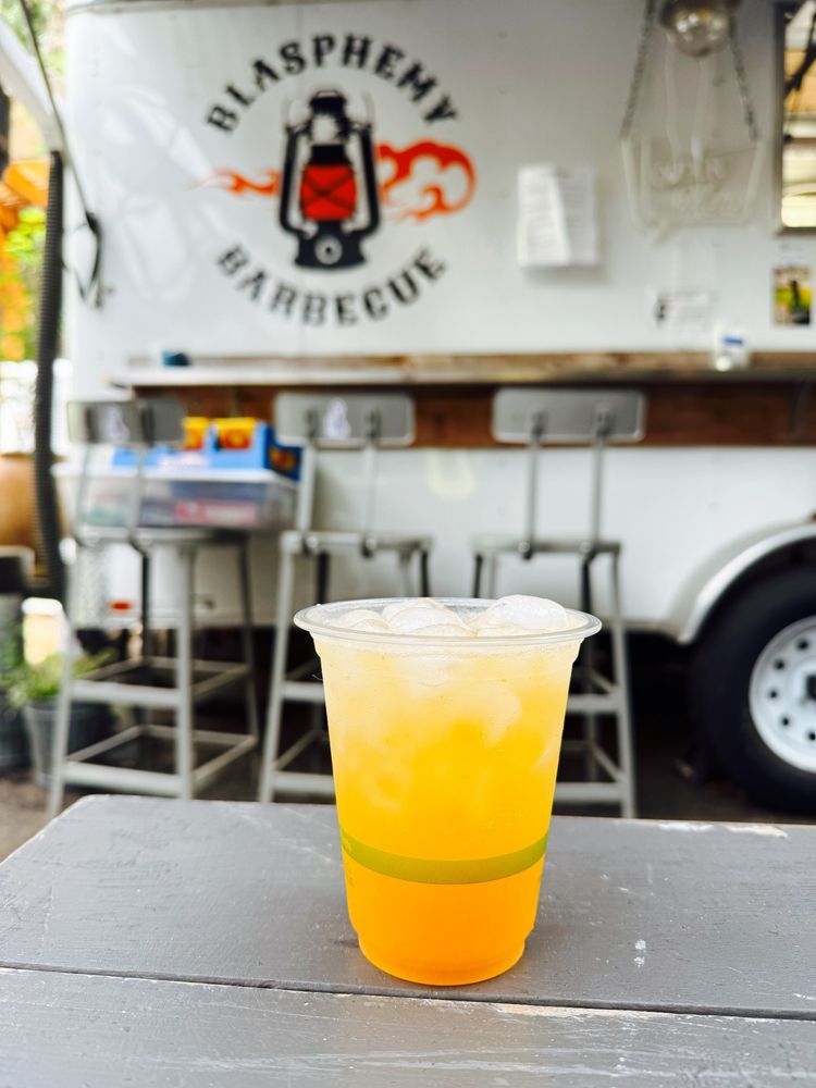 An orange colored cocktail sitting in a picnic table in front of the blasphemy bbq bar cart.