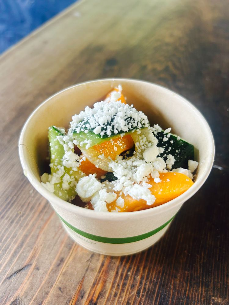A paper bowl with cucumbers, apricots and crumbly white cheese, the  bowl is sitting on a wooden table.