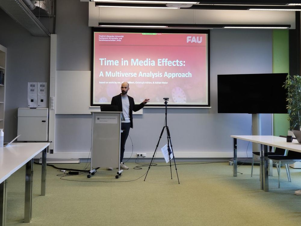 Me standing in a well-lit conference room in front of a presentation titled "Time in Media Effects: A Multiverse Analysis Approach". I wear a black jacket and dark pants with a grey shirt and point at the presentation. 