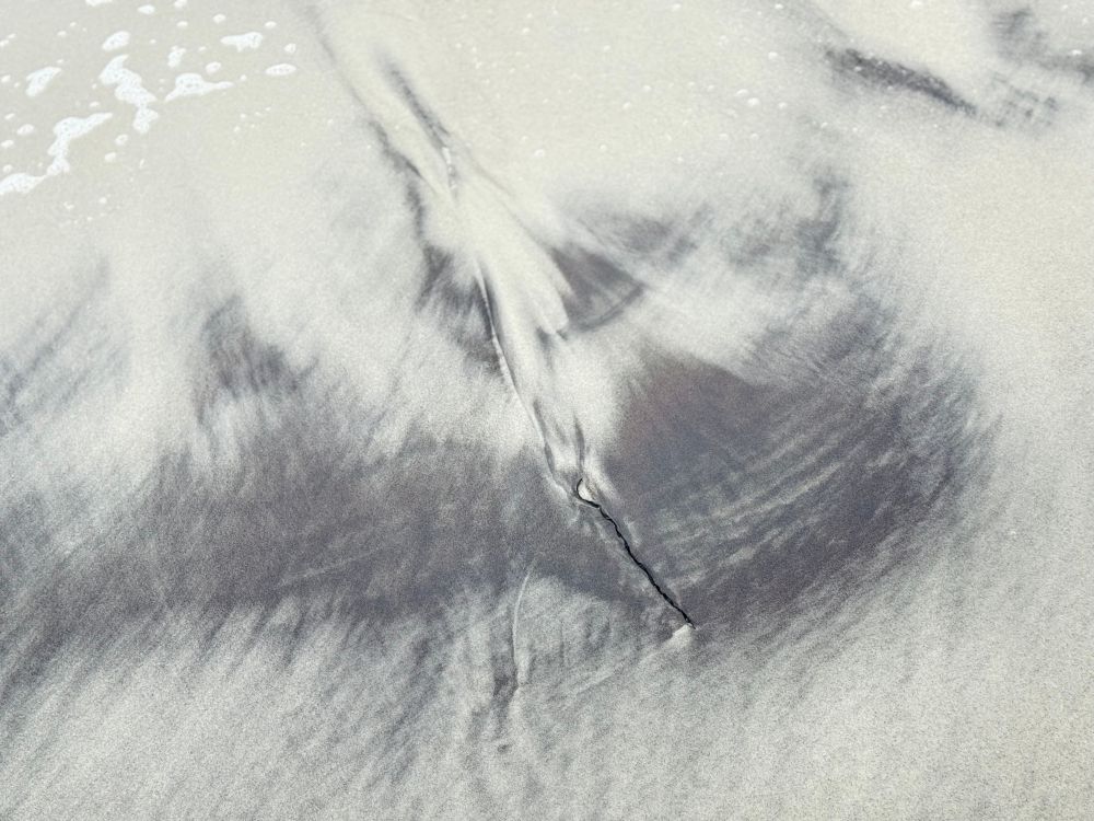 Schwarzer Sand in schönen fließenden Sandschatierungen gemischt in helleren Sand. Durch beide läuft ein kleiner, harter Riss. Links oben im Bild ist Gischt zu sehen. 