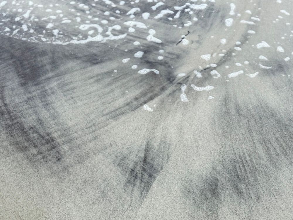 Schwarzer Sand in fächerartigen Sandschatierungen zwischen hellerem Strandsand. Das Meer hat weiße  Gischtflocken darauf verteilt. 