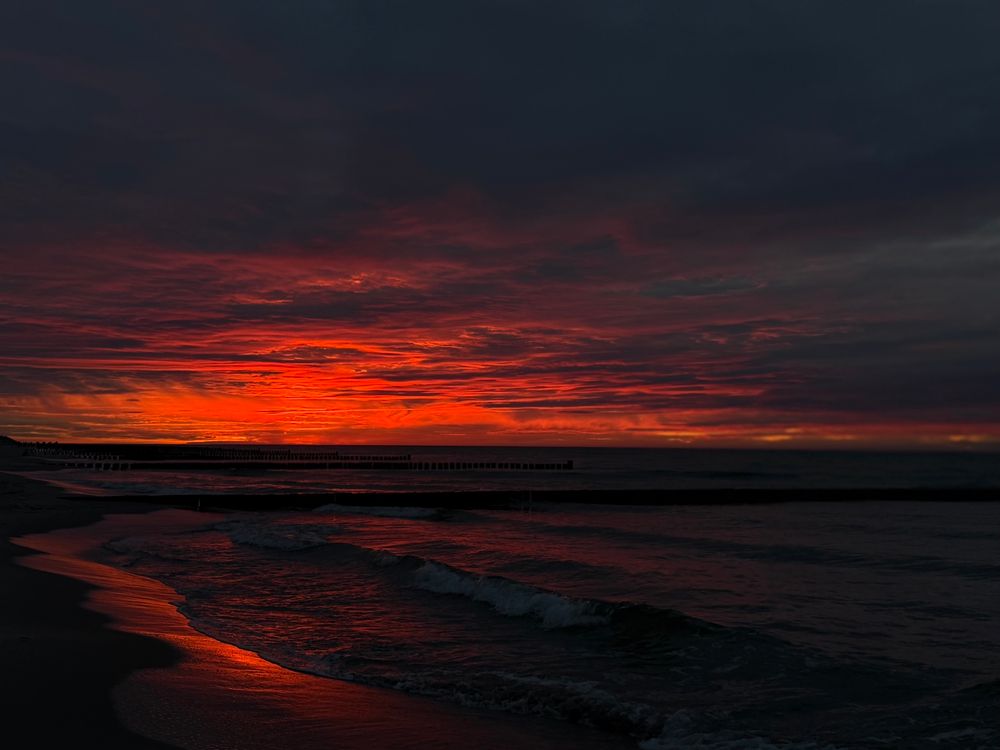 Over a black sea a red and darkblue sky illuminates parts of the sand, the water and the darkness. It is a warm dark red color caused by a solar storm. No filter. 