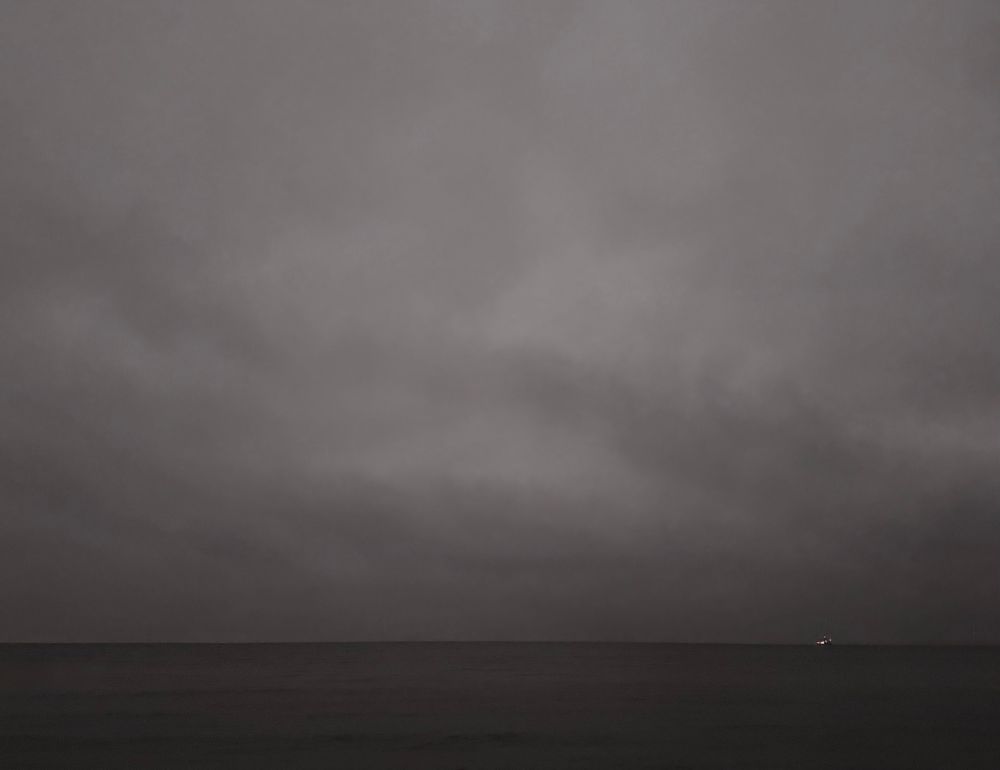 A dark sea and a grey cloudy sky at dusk. A ship crosses Both. It’s lights can be seen from the shore. 