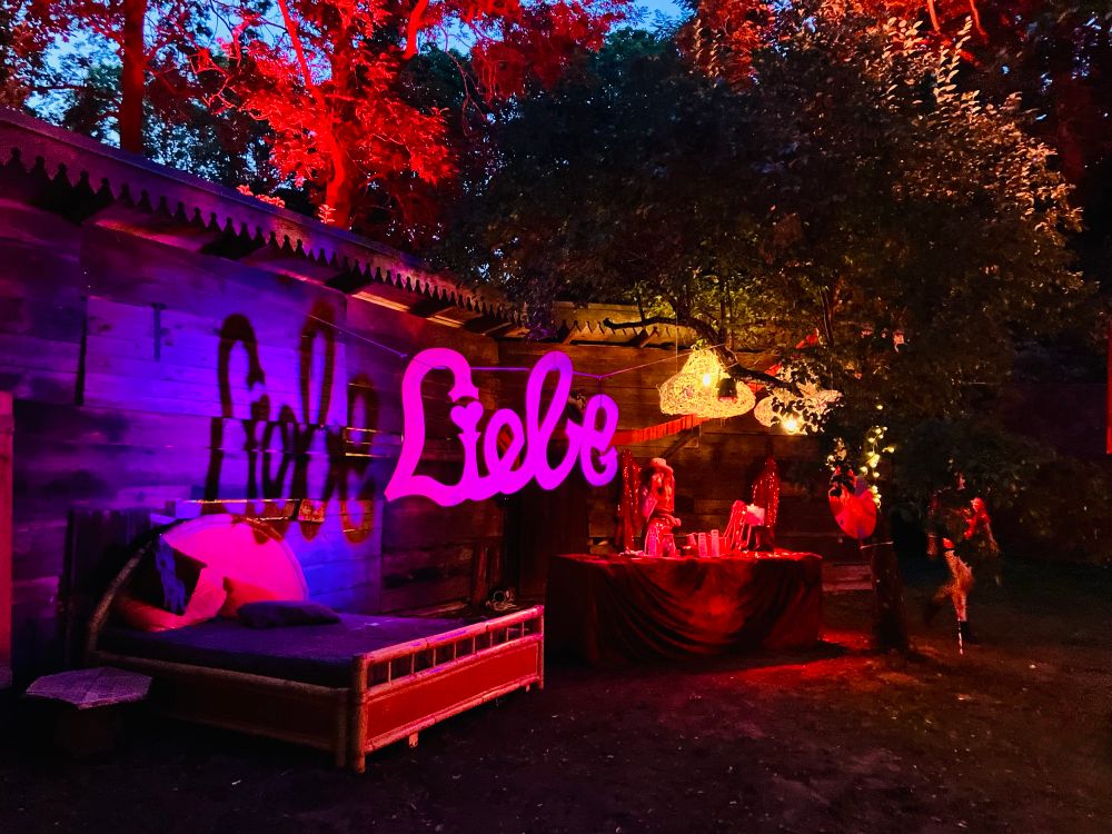 In knalligen bunten rot-, violett- und Blautönen angestrahlter Ort zwischen mehreren großen Laubbäumen. Vor einer HolzbretterWand steht ein HolzDoppelbett mit bunten Kissen und Decken. Rechts daneben ist eine kleine Bar aufgebaut, über der zwei große goldgelb leuchtende Lampen hängen. Zwischen Bett und Bar schwebt ein knallpinkfarbener Schriftzug „Liebe“. Hinter der Bar steht eine Frau mit Hut, die etwas trinkt. Rechts von der Bar läuft jemand an einem Laubbaum vorbei. Durch die großen Laubbäume blitzt noch etwas blau des langsam dunkel werdenden Himmels hindurch. s