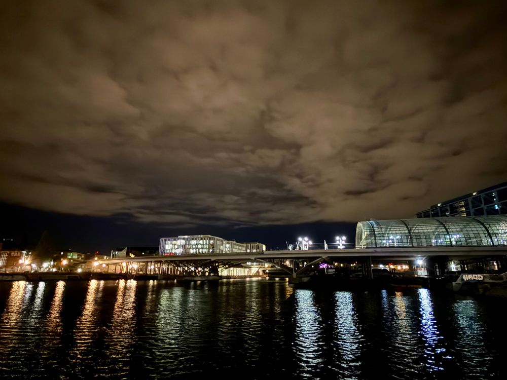 Blick auf den Humboldthafen und die Humboldtbrücke bei Nacht. Das Licht verschiedener Gebäude und Laternen spiegelt sich in unterschiedlichen Farbschattierungen im Wasser, auf dem leichte Wellenbewegungen zu sehen sind. Der Himmel ist dicht bewölkt.