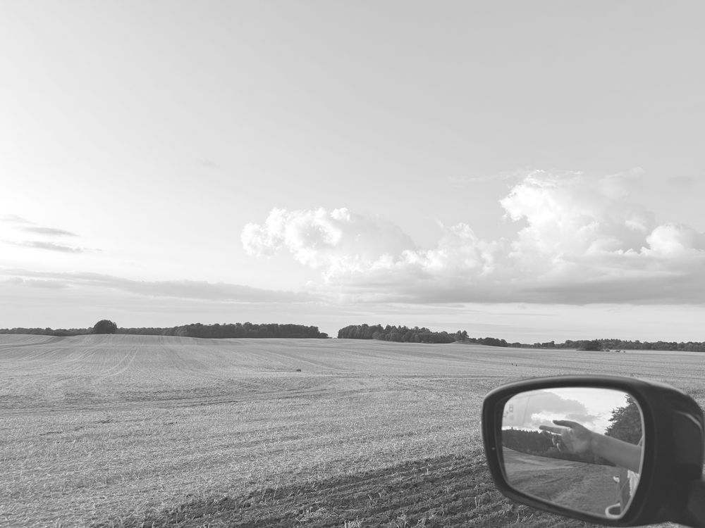 Schwarz-weiß Aufnahme eines Ackers aus einem Autofenster. Am Himmel hängt tief eine einzelne große weiße Wolke, in der Ferne an der Ackerkante ist Wald zu erkennen. Im Seitenspiegel sieht man Acker, Wald und Himmel und eine linke Hand, die aus dem Fenster entspannt in den Fahrtwind gehalten wird. 