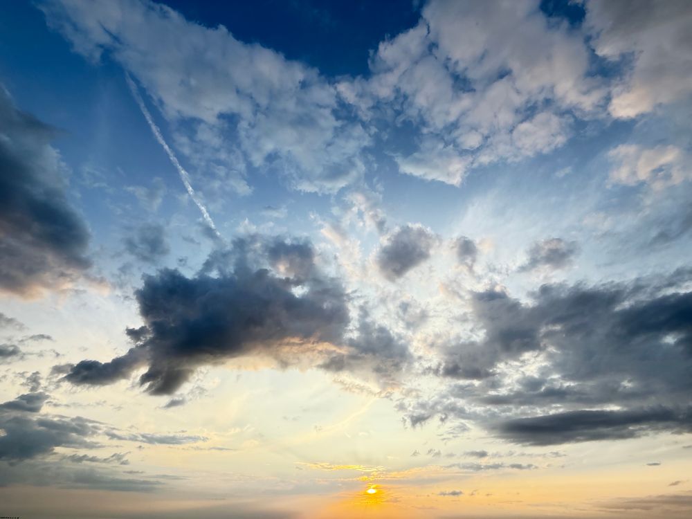 Im Westen tiefstehende Sonne als warmorangefarbener kleiner Ball am Himmel über dem Meer, das nicht im Bild zu sehen ist. Von Osten ziehen dunkle Wolken Richtung Sonne. Kein Filter. 
