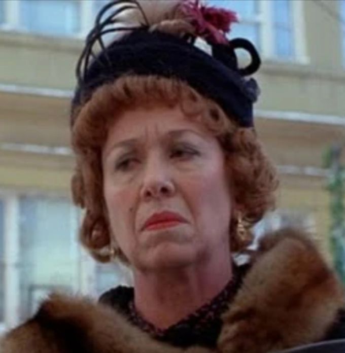 An old woman wearing a hat and a fur stole over her shoulders. She has a mean look on her face.