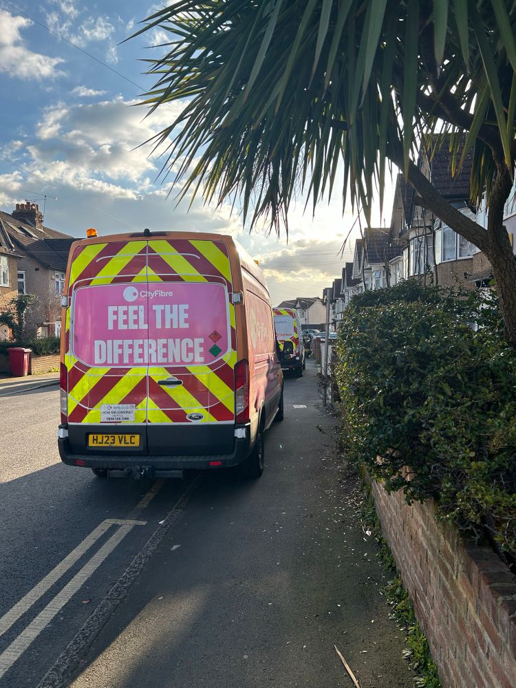 A van parked on the pavement, blocking it for wheelchairs and buggies.
The van has a sign asking How am I driving? with a phone number to call on the back.