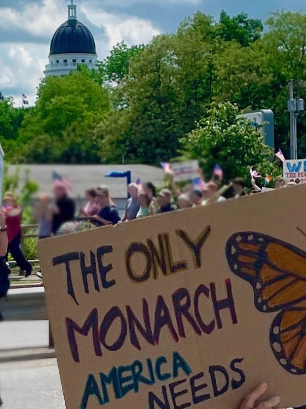 In the foreground, a sign pained on cardboard that says The Only Monarch America Needs with a drawing of a monarch butterfly. In the mid ground, some marchers carrying American flags. In the background, the dome of the state capitol is visible.