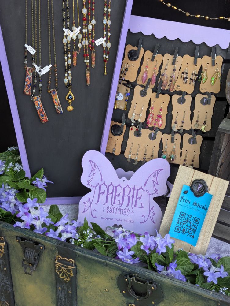 the interior of the chest filled with jewelry on display. a lilac colored sign in the shape of a butterfly reads "faerie earrings individually priced". a second blue sign reads "Fehu Studio", the name of the artist who created all these treasures in the chest.