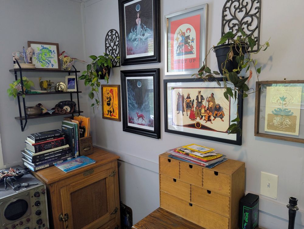 the most pleasant corner of my otherwise grey office. a shelf with knickknacks and various prints hang above books and zones stacked on wood furniture. two vining plants in iron planters frame the wall on either side