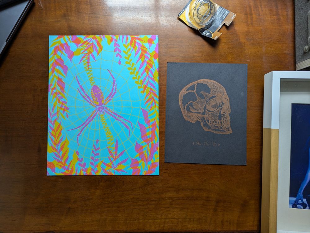 a screen print of a black and yellow orb weaver on teal paper and a screen and letterpress print skull on black paper with text reading "ain't dead yet"