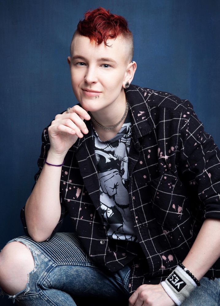 My headshot! I has a short red fauxhawk and I be smiling. I’m wearing a plaid black shirt over a black and white marvel comics tee and jeans.
