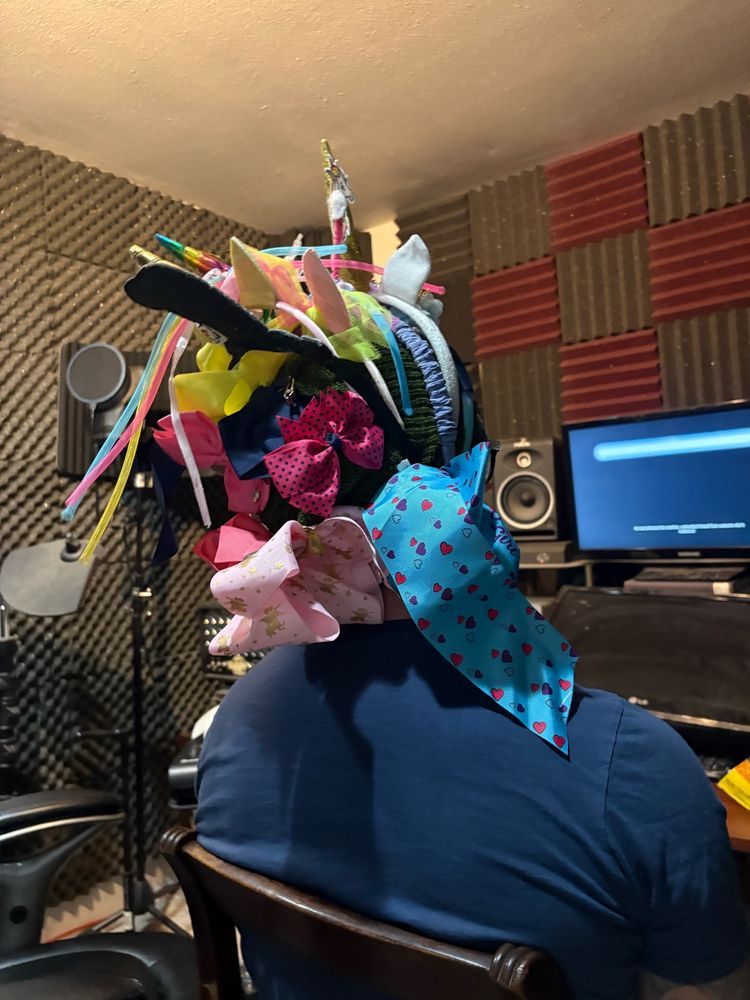 A man sitting in a chair with many hair bows and head bands attached to his beanie 