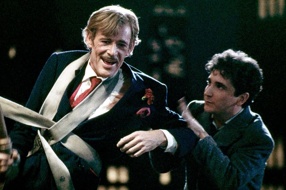 An older Peter O'Toole dressed in a fancy suit with a firehose wrapped around his chest while weaving around drunkenly. A young man in a shabby suit tries to control him. 