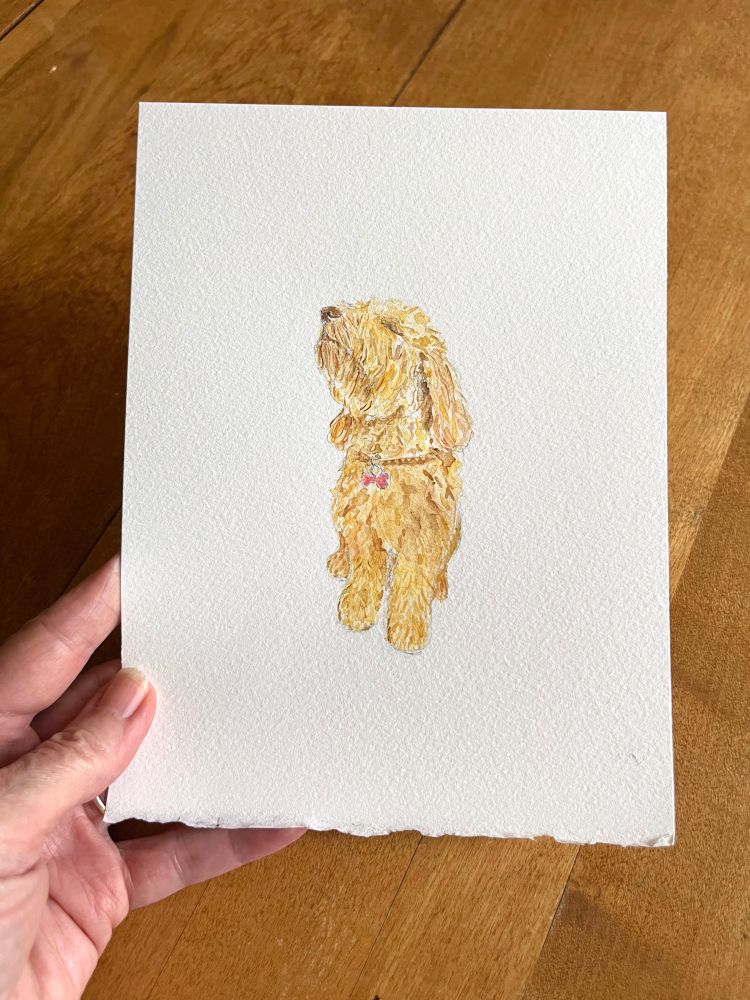 My hand holding a watercolour painting I did of my goldendoodle Jasper. In the painting, he has his head tilted back and his nose in the air in a moment of pure bliss. Artist: Kathleen Maunder