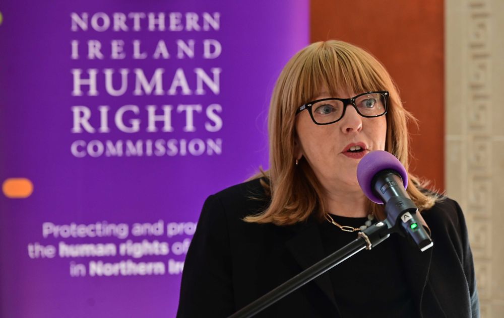 Alyson Kilpatrick in foreground speaking into a microphone, NIHRC purple banner behind her