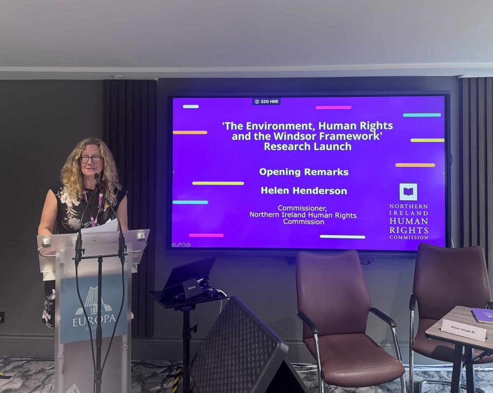 NIHRC Commissioner Helen Henderson speaking from a podium in front of a purple holding slide.