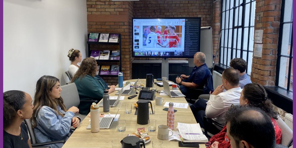Nine people sitting in a boardroom. A presentation can be seen on a television screen that reads UEFA Foundation for Children. NI and Human Rights Forum Meeting. 