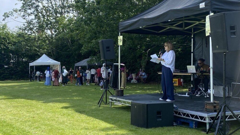 Small stage and gazebo on grass with a speaker holding a microphone, people standing in the background