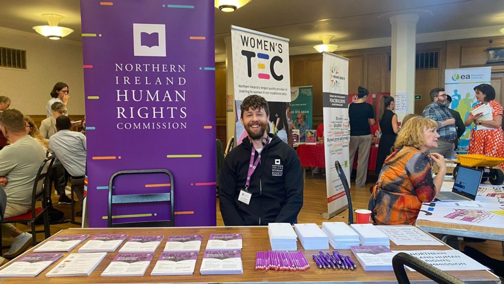 A NIHRC staff member at the Big Sanctuary Information Fair. He is sitting at a table, in front of a large purple pop up banner that reads "Northern Ireland Human Rights Commission".