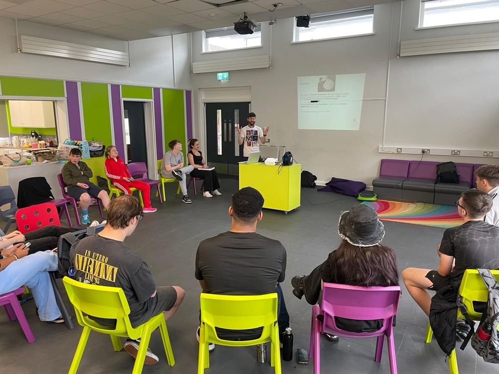 Young people sit on brightly coloured chairs in a circle, with a person giving a presentation from the front