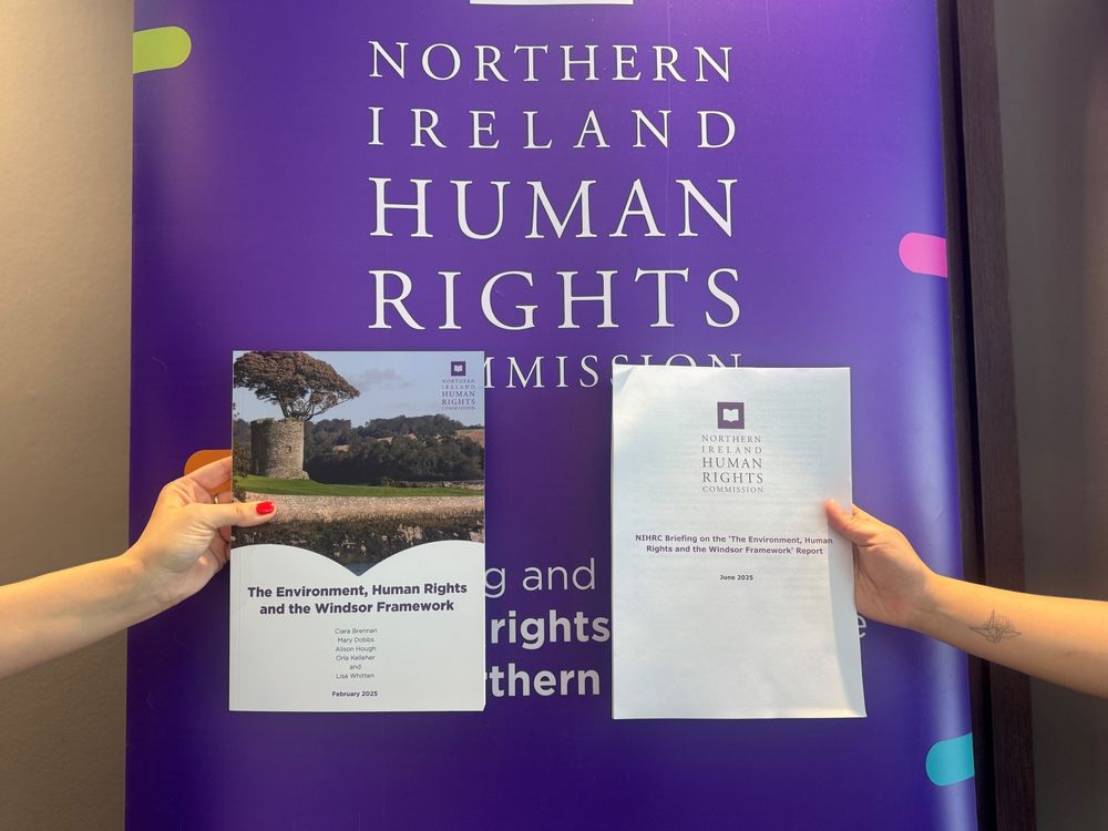 A photo of two hands in front of a purple pop up banner reading "Northern Ireland Human Rights Commission". One holds a copy of the 'The Environment, Human Rights and the Windsor Framework", and the other holds a copy of the NIHRC briefing paper.