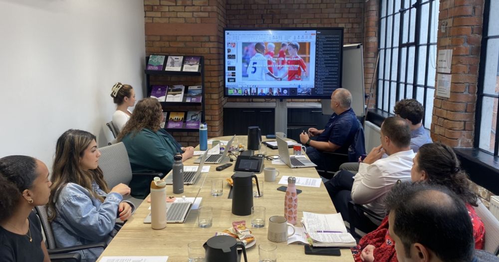 People sit around a boardroom table, looking away from the camera at a screen.