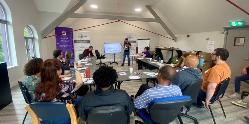 People sitting in a U shaped table. At the front, NIHRC staff are presenting the work of the Commission. Three pop-up banners can be seen: one with the NIHRC logo and two with the Migrant Centre NI logo.