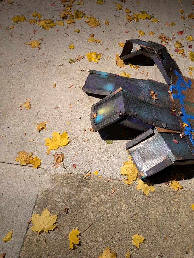 Blocky looking fingers have been shaped out of cardboard and built up to have depth.  Each finger is about 2 feet long and attached to a flat cardboard "palm" that is only partially in frame.  The fingers have been painted with spay paint and have a dark background with splotches of other colors. Blue masking tape lines the edges of the palm. The part of the palm visible is not painted.