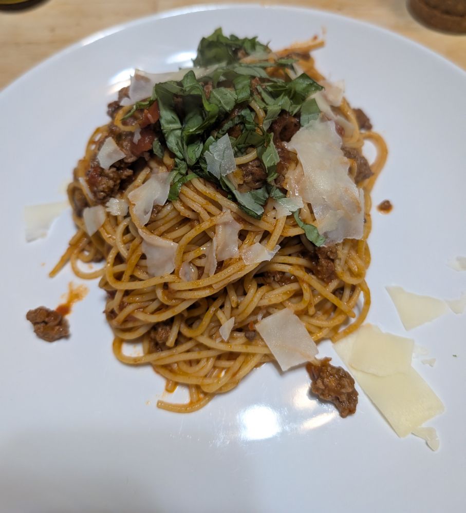 On a white dinner plate: noodles that have been finished with a meat sauce that also contains sliced mushrooms.  Topped as described with shaved parmesan and sliced basil.  