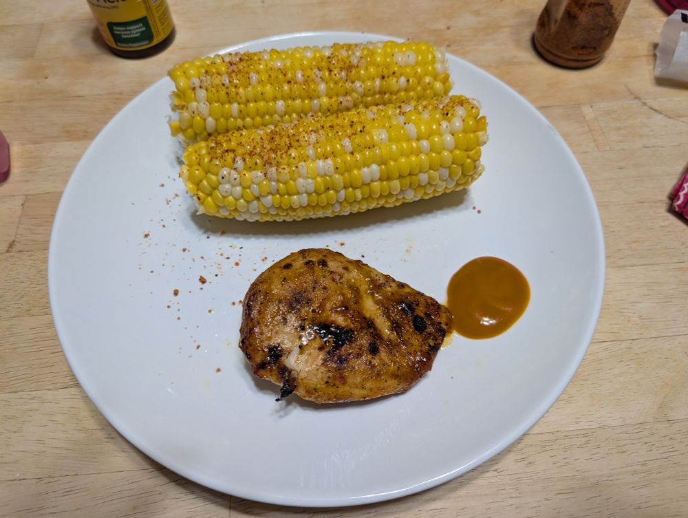 On a white dinner plate: two cobs of corn that have been buttered and sprinkled with tajin, a spicy and lemony Mexican seasoning. A small portion of bbq chicken breast is also on the plate, a bit dwarfed by the corn. There is a dollop of Carolina gold bbq sauce next to the chicken.  Behind the plate on the table, the bottoms of the bbq sauce bottle and the tajin bottle are at the top of the picture. 