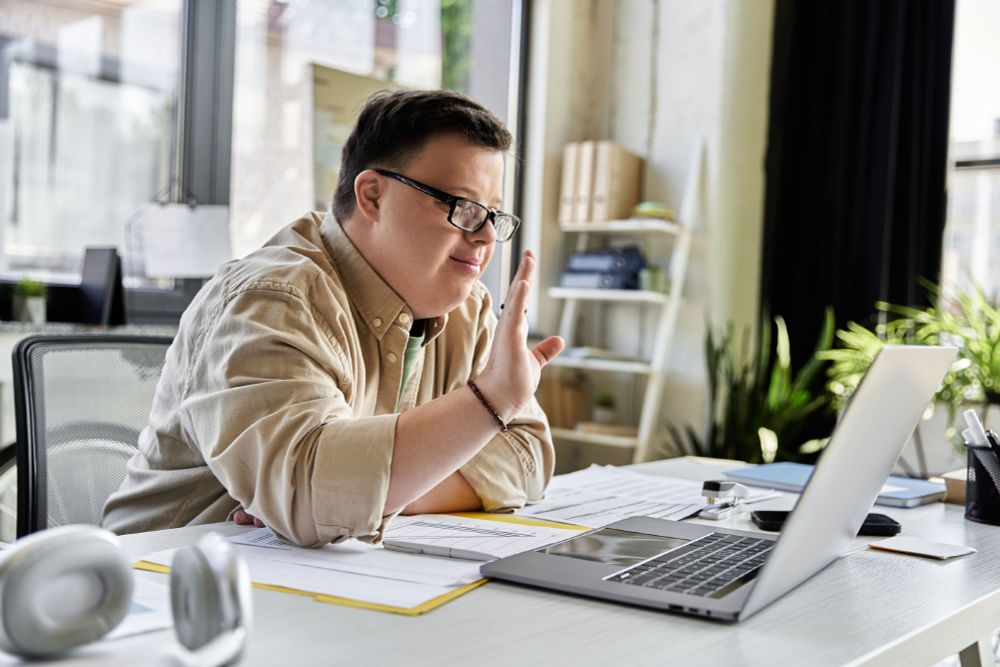 man with Down syndrome, with brown hair, glasses, and long-sleeve beige shirt, waves to someone on his laptop
