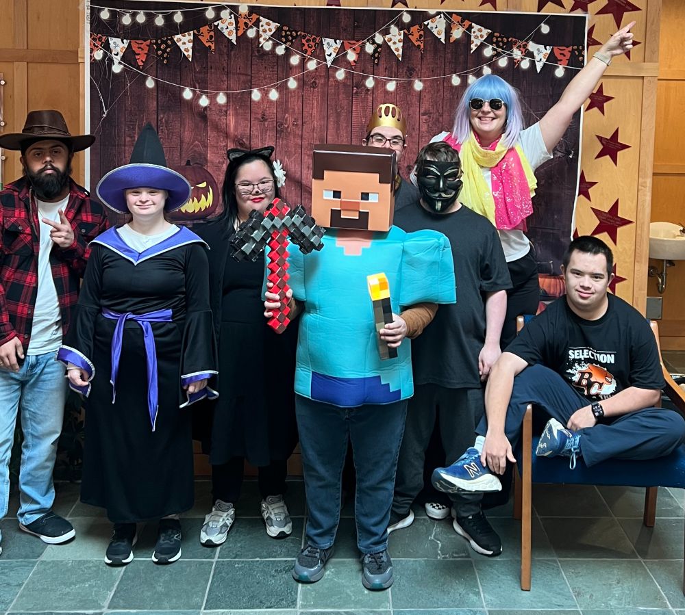 a group of adults with Down syndrome dressed for Halloween stand together in front of a spooky backdrop