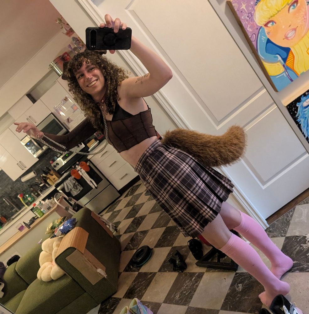 a mirror photo of ari. she's wearing a brown and pink plaid skirt, pink knee high socks, a black bra, a brown shirt that has one sleeve and goes under her armpit on the other side, a fluffy brown tail, brown dog ears, and a collar with a silver chain attached. she's holding the chain in one outstretched arm and taking the photo with her other hand. 
she's a puppy basically 