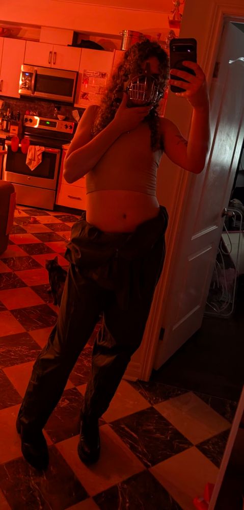 mirror selfie photo of me in red lighting,, in a house,,
im wearing shiny black boots, a green flight suit rolled down to my hips, a crop top, and I'm holding a dog muzzle on my face